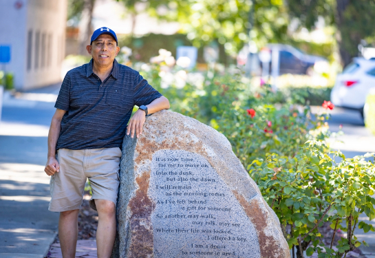 Man with dark skin wearing a blue hat leaning his left arm on a large rock with writing on it.